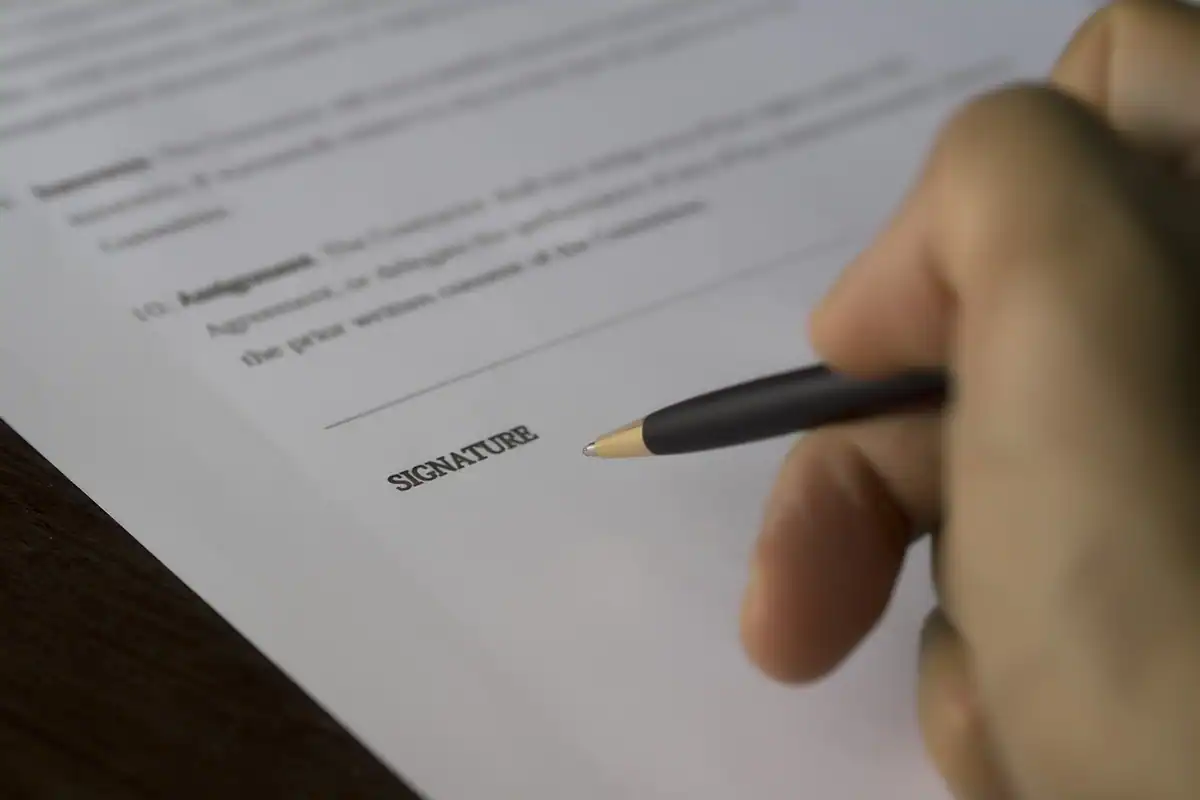 O mână semnează cu pixul un contract tipărit, așezat pe masă