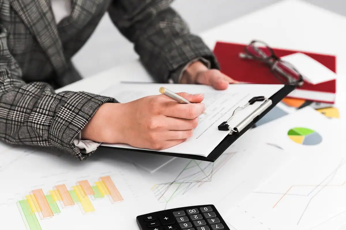 O mână completează un formular pe clipboard, lângă grafice și un calculator