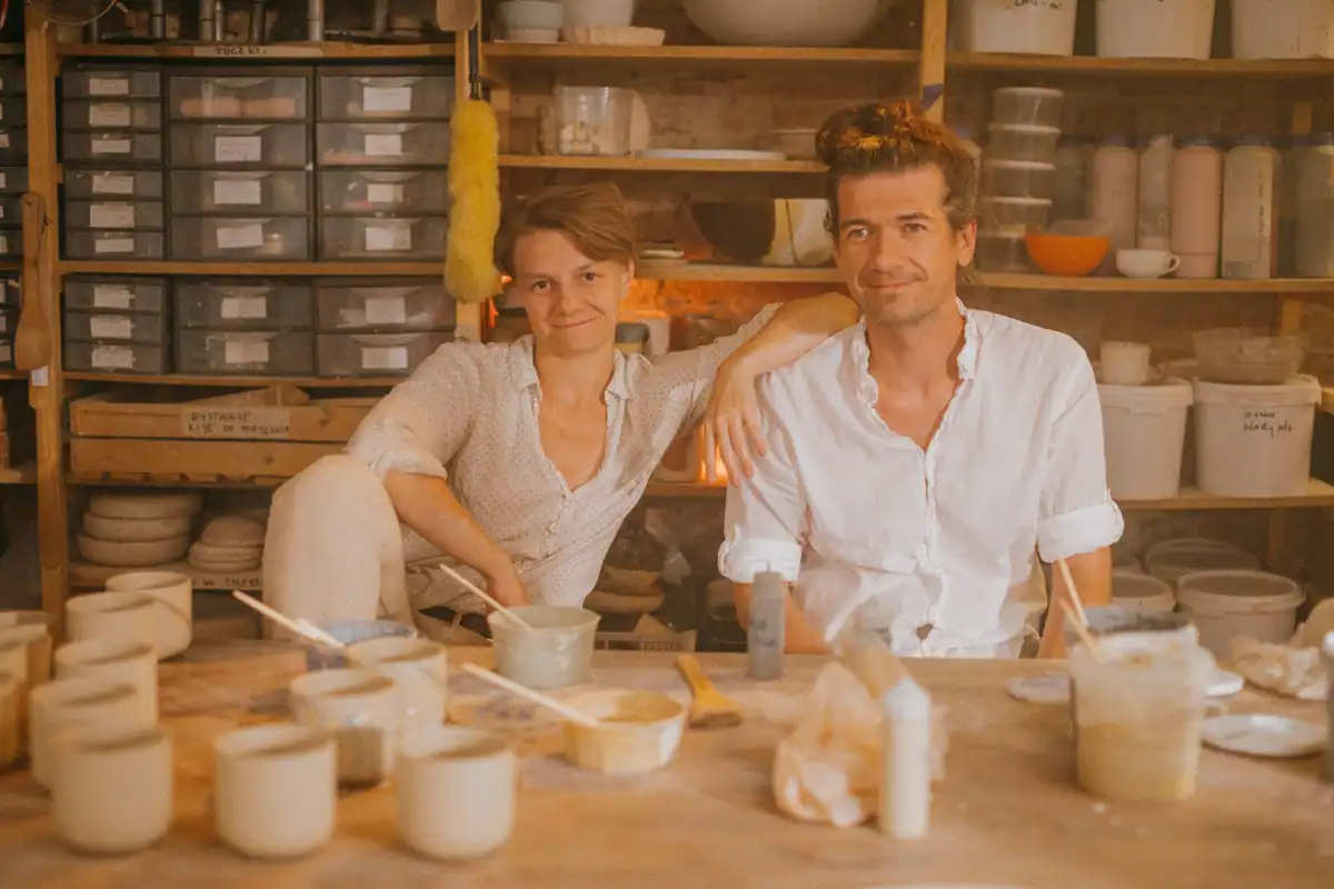 Doi parteneri zâmbesc într-un atelier cu rafturi și castroane pe masă